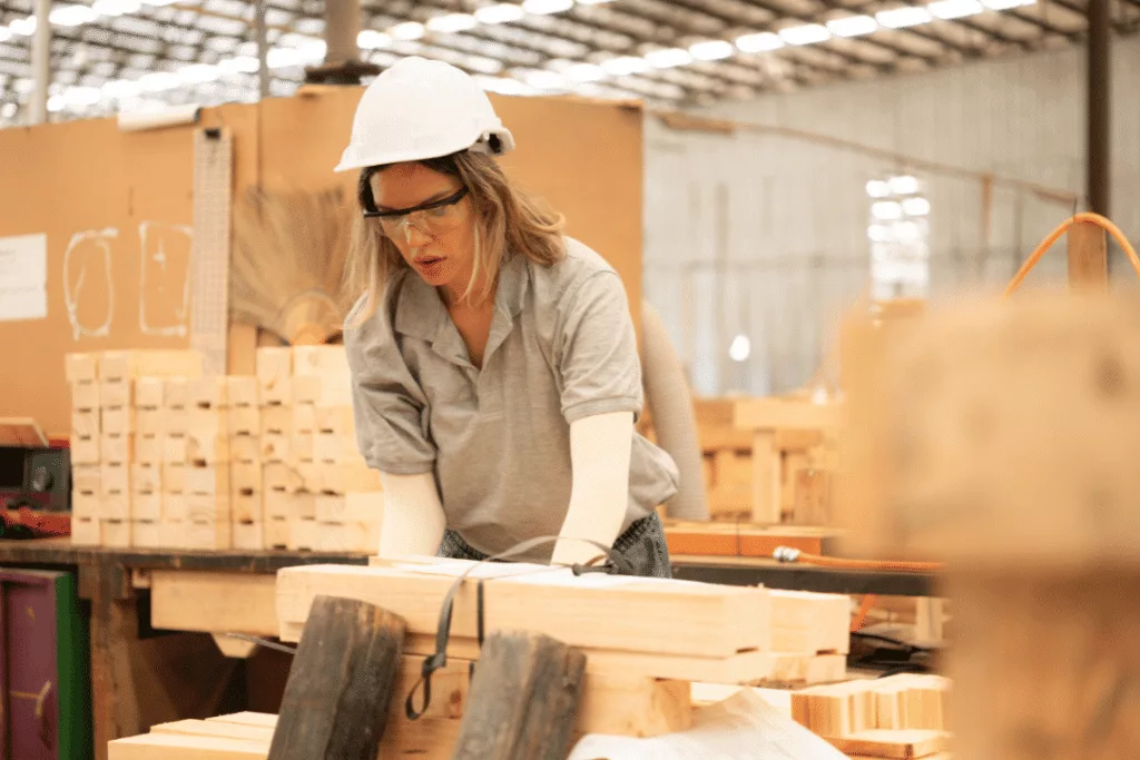 A person wearing a hard hat, safety glasses, and gloves works with wooden planks and a pallet in an industrial workshop setting.