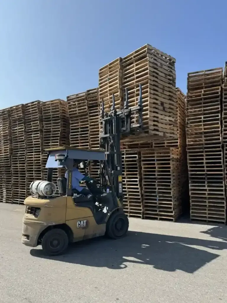 A forklift operator lifts a stack of wooden pallets in front of large pallet piles under a clear blue sky, showcasing efficient handling often seen at pallet recycling near me facilities.